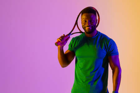 Image of happy african american male tennis player in violet and yellow neon lighting. Sport, movement, fitness and active lifestyle concept.の写真素材