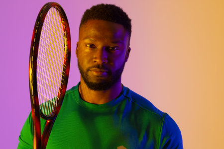 Image of african american male tennis player in violet and yellow neon lighting. Sport, movement,...