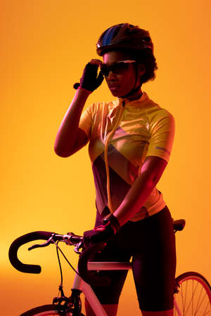 Vertical image of african american female cyclist with bike in neon orange lighting. Sport, movement, fitness and active lifestyle concept.の写真素材