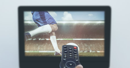 Composition of hand of sports fan holding remote control, with football match on tv screen. sport...