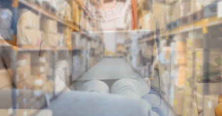 Mid section of a Caucasian woman using laptop computer at home shopping on line over warehouse with shelves full of cardboard parcel boxes in the background. Online shopping ecommerce social distancing and self isolation in quarantine lockdown concept digital composite.の写真素材