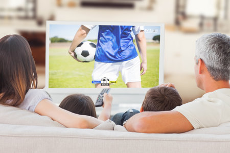 Family sitting on sofa watching television together against football players tacakling for the ball on pitchの写真素材