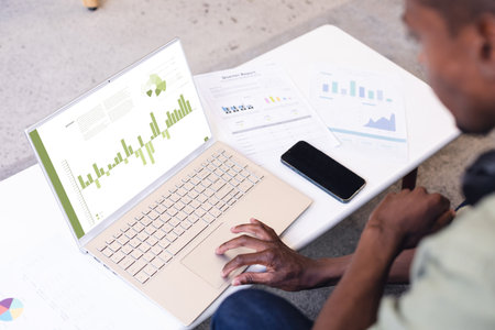 African american male professional with documents and cellphone on desk analyzing report over laptop. Unaltered, office, entrepreneur, finance, investment, data, performance and technology concept.の写真素材