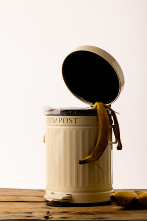 Vertical image of banana skin hanging out of open kitchen composting bin. On white background with...