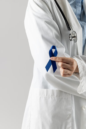 Midsection of caucasian doctor holding blue ribbon on grey background. Medicine, healthcare and science concept.の写真素材