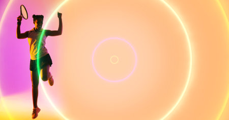 Full length of cheerful biracial female tennis player with racket dancing by illuminated circles. Copy space, composite, sport, competition, playing, match, winning, shape, glowing and abstract.の写真素材