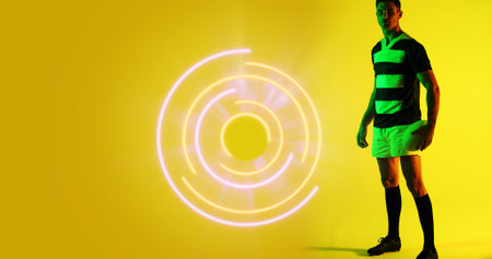 Full length of caucasian rugby player with ball standing by illuminated circular pattern, copy space. Yellow, composite, sport, competition, playing, match, shape and abstract concept.の写真素材