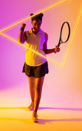 Happy african american female tennis player holding racket and shaking fist by glowing triangle. Copy space, composite, shape, celebration sport, competition, winner, match and abstract concept.の写真素材