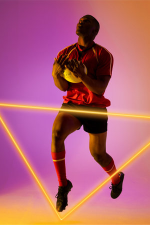Full length of african american male rugby player catching ball and jumping by illuminated triangle....