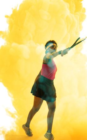 Full length of african american female player hitting with racket over yellow smoky background. Copy...