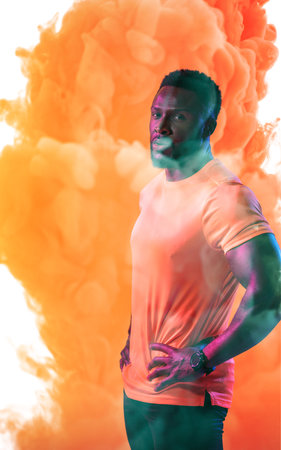 Portrait of confident male athlete with hands on hip standing against orange smoky background. Copy...