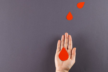 Hand of caucasian woman holding blood drop, with blood drops on grey background, copy space. Blood donation, medicine and healthcare.の写真素材