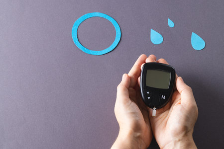 Hands of caucasian woman holding glucometer over blue drops and ring on grey background, copy space. Blood sugar, diabetes and health awareness.の写真素材