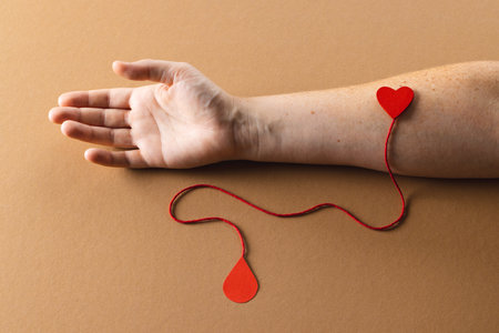 Arm of caucasian woman with heart and red string with blood drop, on brown background. Blood donation, medicine and healthcare.の写真素材