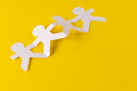 Close up of four paper cut out people figures holding hands with copy space on yellow background. Humanitarian, people, help and human concept.の写真素材
