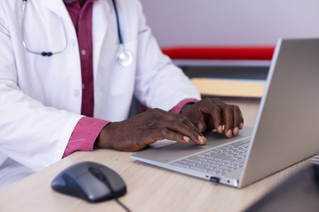 Midsection of african american male doctor sitting at desk using laptop at doctor&#39;s office. Hospital, medicine, healthcare and communication.の写真素材
