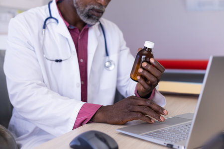 African american male doctor sitting at desk holding medicine and using laptop at doctor&#39;s office. Hospital, medicine, healthcare and communication.の写真素材