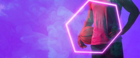 Midsection of biracial basketball player with ball standing by hexagon over smoky background. Blue, composite, copy space, hand, sport, competition, illustration, illuminated, shape and abstract.の写真素材