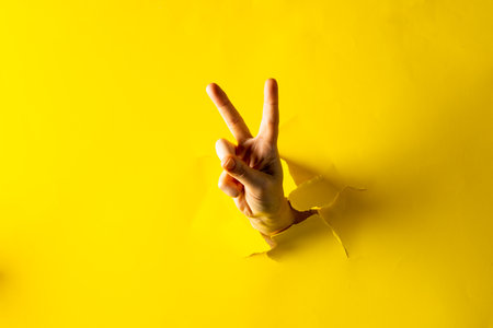 Close up of hand of caucasian woman showing peace sign with copy space on torn yellow background. Peace and anti war movement concept.の写真素材