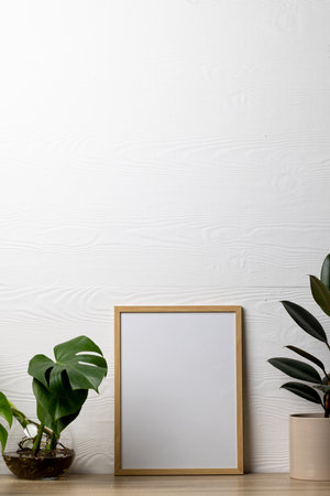 Vertical of empty wooden frame with copy space and plants in pots on table against white wall. Mock...