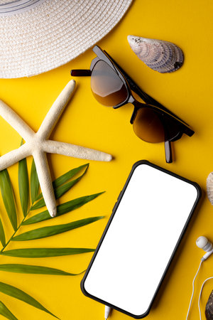Smartphone with copy space on screen, straw hat, sunglasses, plant, seashells on yellow background....