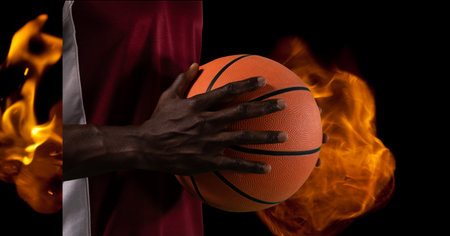 Composition of midsection of male basketball player holding ball over flames on black background. sport and competition concept digitally generated image.の写真素材
