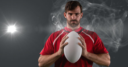 Portrait of caucasian male rugby player holding ball against smoke and light spot on grey background. sports tournament and competition conceptの写真素材