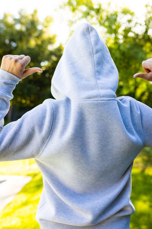 Rear view of caucasian woman pointing fingers and wearing gray hoodie over green park on sunny day....