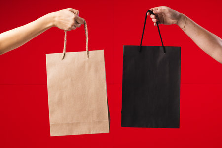Hands of caucasian women holding black and brown gift bags with copy space on red background. Black friday, cyber monday, shopping, cyber shopping, sales, retail and shipping concept.の写真素材