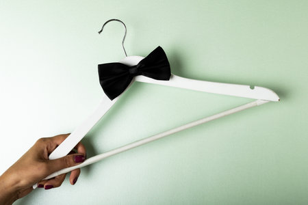 Cropped hand of biracial woman holding hanger with black bowtie over blue background, copy space. Menswear, fashion, elegance concept.の写真素材