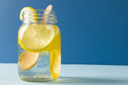 Close-up of mason jar with lemonade and lemon slices on table against blue background, copy space....