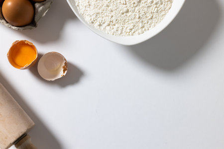 Directly above shot of flour in bowl with broken egg and rolling pin on white background, copy space. food, cookie, ingredients and preparation concept.の写真素材