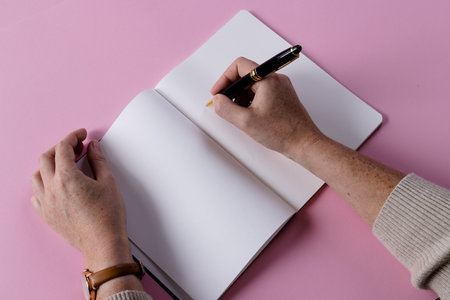 Hands of caucasian woman holding pen and writing in notebook with copy space on pink background. Literature, writing, leisure time and books.の写真素材