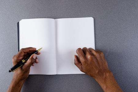 Hands of biracial man holding pen and writing in notebook with copy space on grey background. Literature, writing, leisure time and books.の写真素材