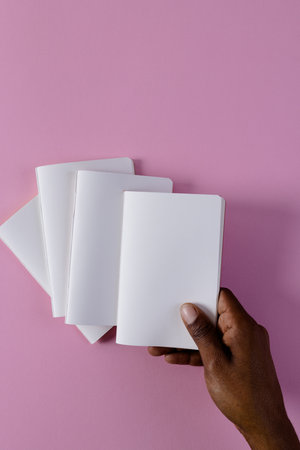 Hand of biracial man holding notebook over notebooks with copy space on pink background. Literature, reading, leisure time and books.の写真素材