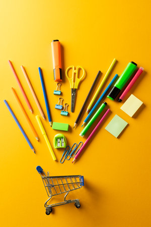 Close up of miniature shopping trolley with school materials on orange background. Back to school, shopping, school materials, learning, school and education concept.の写真素材