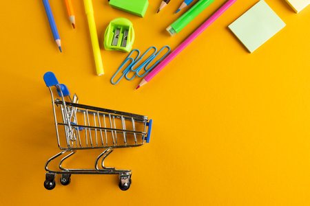 Close up of miniature shopping trolley with school materials, copy space on orange background. Back to school, shopping, school materials, learning, school and education concept.の写真素材