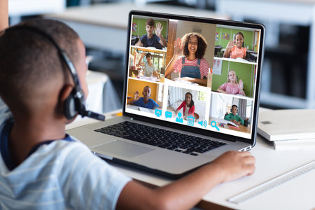 African american boy looking at students and teacher waving hands over laptop during online class....