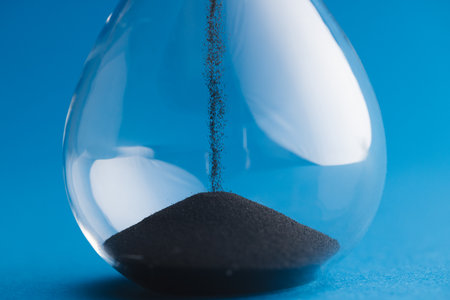 Close up of hourglass with black sand and copy space on blue background. Time, timekeeping, shape...