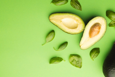 Close up of two halves of avocado and basil leaves and copy space on green background. Vegetable,...