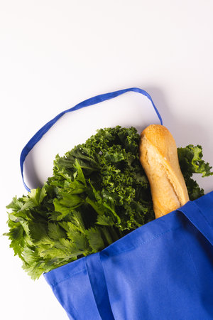 Blue canvas bag with baguette and green salad vegetables and copy space on white background. Fresh food, shopping, bag, colour, fabric, texture and materials concept.の写真素材