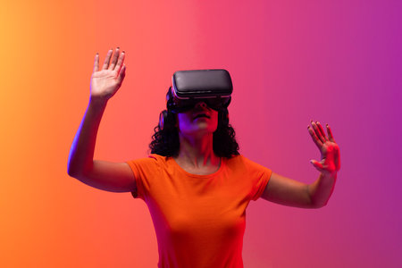 Biracial woman using vr headset and pointing on neon pink to orange background. Technology, digital networks, global connections and communication metaverse.の写真素材