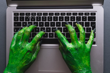 Green monster hands writing on laptop keyboard on grey background. Halloween, tradition and...