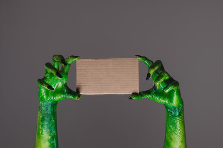 Green monster hands holding cardboard with copy space on grey background. Halloween, tradition and...