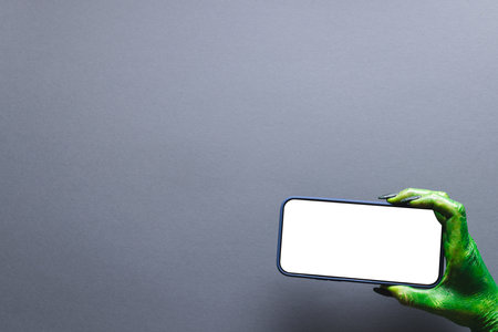Green monster hand holding smartphone with copy space on grey background. Halloween, tradition and...