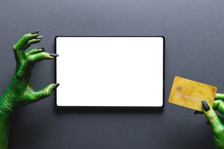 Green monster hands holding credit card and tablet with copy space on grey background. Halloween, tradition and celebration concept.の写真素材