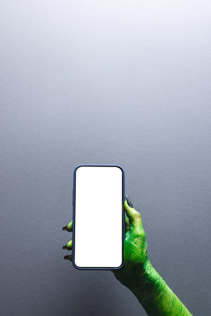 Vertical image of green monster hand holding smartphone with copy space on grey background....
