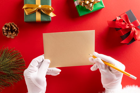 Santa claus holding envelope and pencil with copy space and christmas presents on red background....