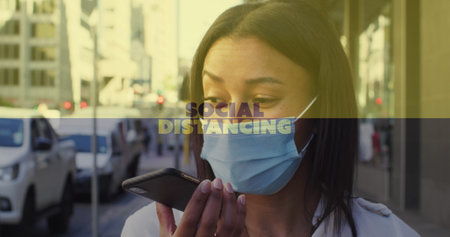 Social distancing text against woman wearing face mask talking on smartphone on the street. global...