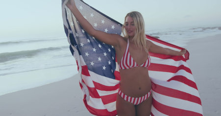 Happy 4th of july text against caucasian woman wrapping american around her on the beach. american...